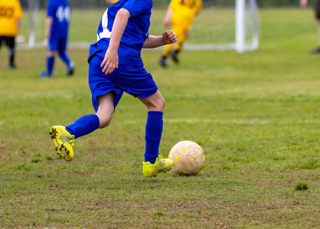 Young boy wearing a blue jersey is kicking a soccer ball on a soccer field